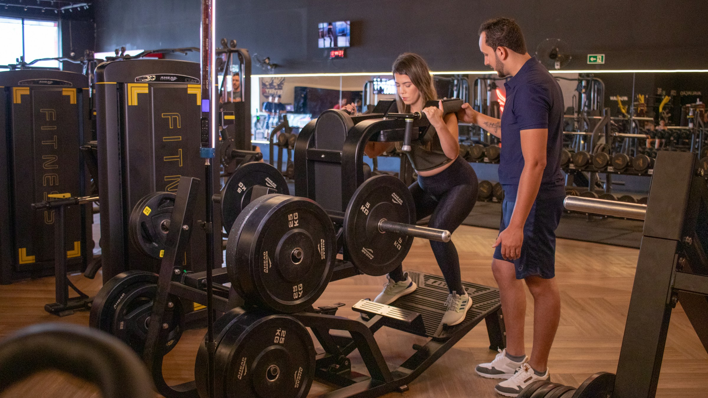 A woman performing a squat on a plate-loaded machine while a trainer watches her form in a gym