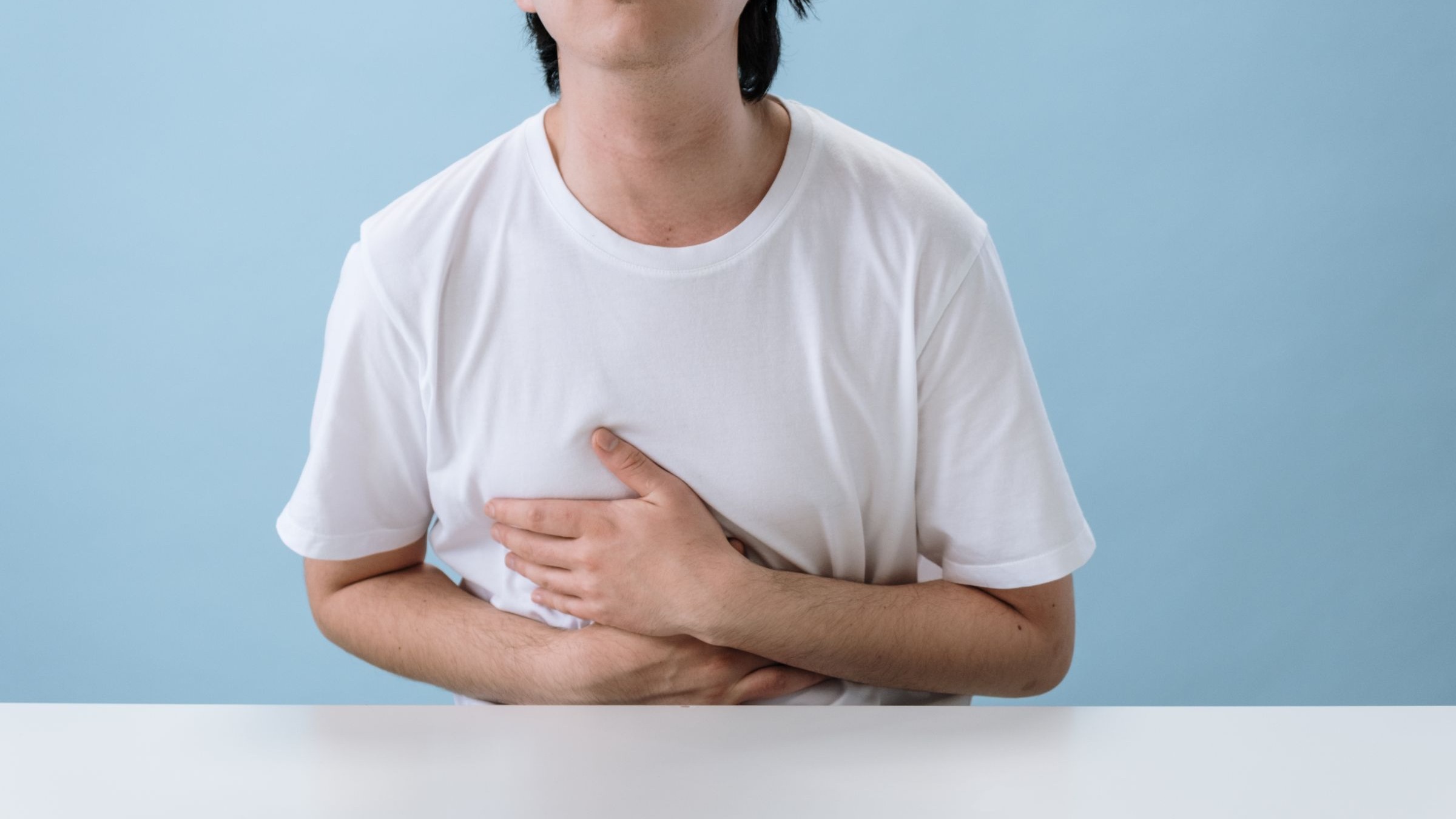 A man in a white shirt holding his abdomen against a light-blue background