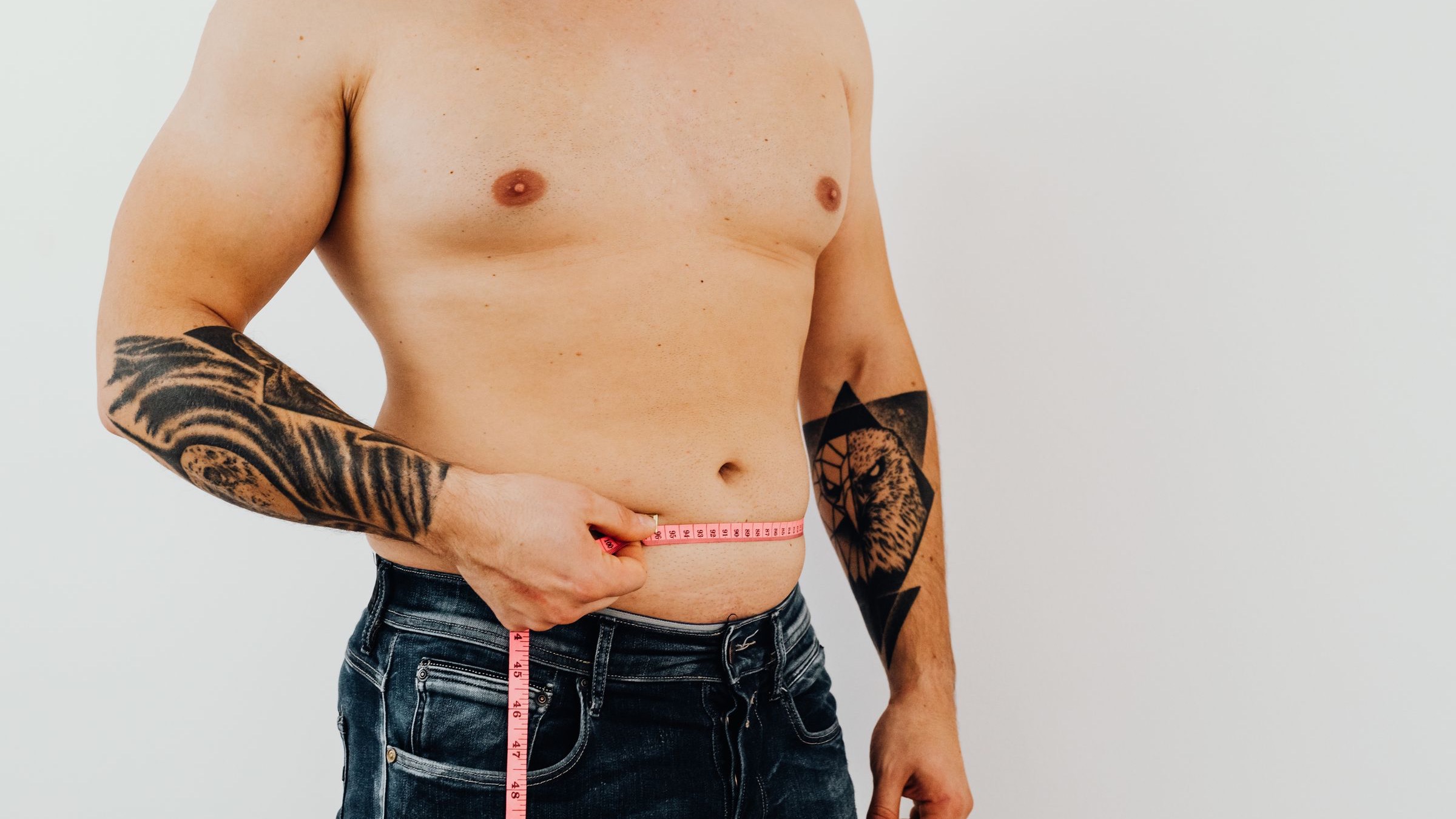 A shirtless man measures his waist with a pink tape measure against a light gray background