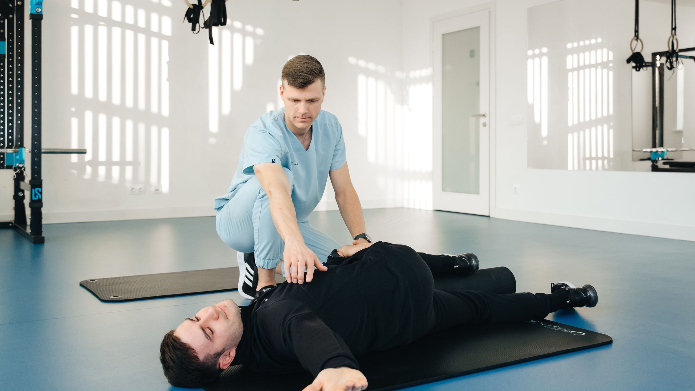 A physiotherapist supports a client performing a floor-based rehabilitation exercise in a bright clinic